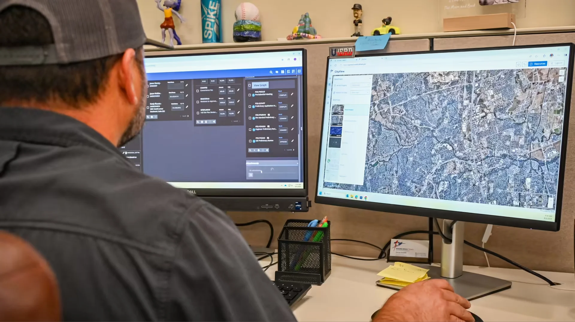 Person wearing a cap works at a desk with two monitors; one shows a control panel interface, and the other displays a detailed satellite map.
