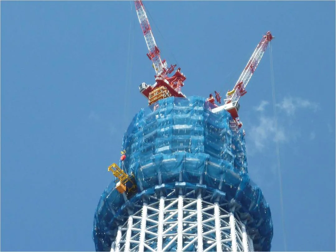 Close-up of the Tokyo Skytree under construction, with cranes at the top and blue netting covering the structure.