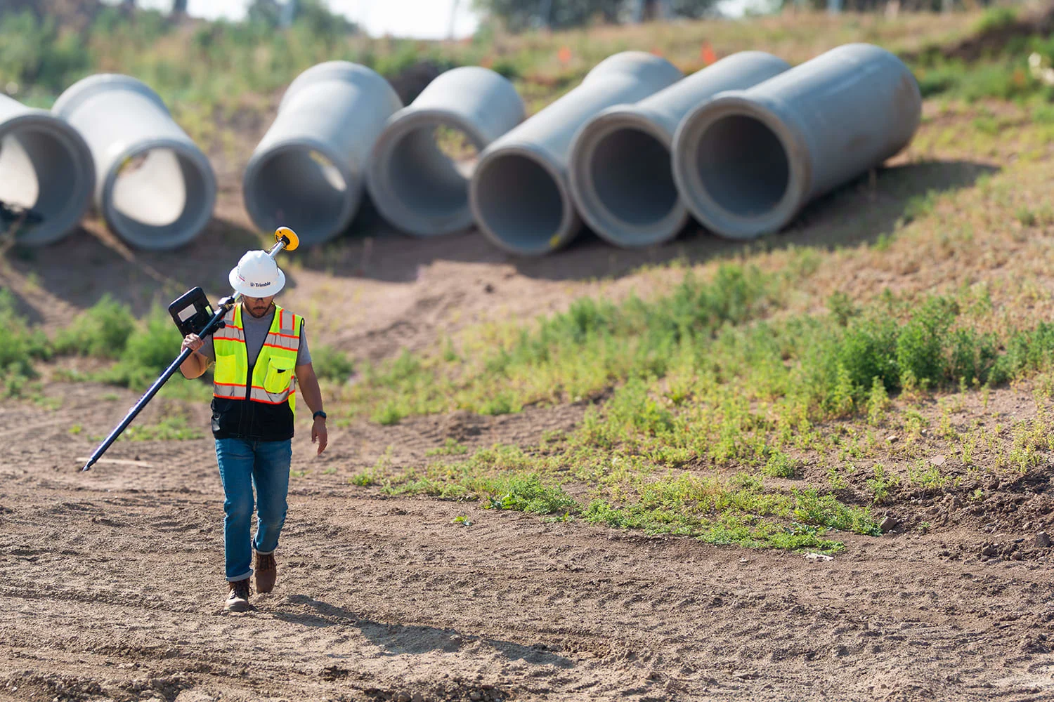 A surveyor walks on a construction site carrying surveying equipment, with concrete pipes in the background.