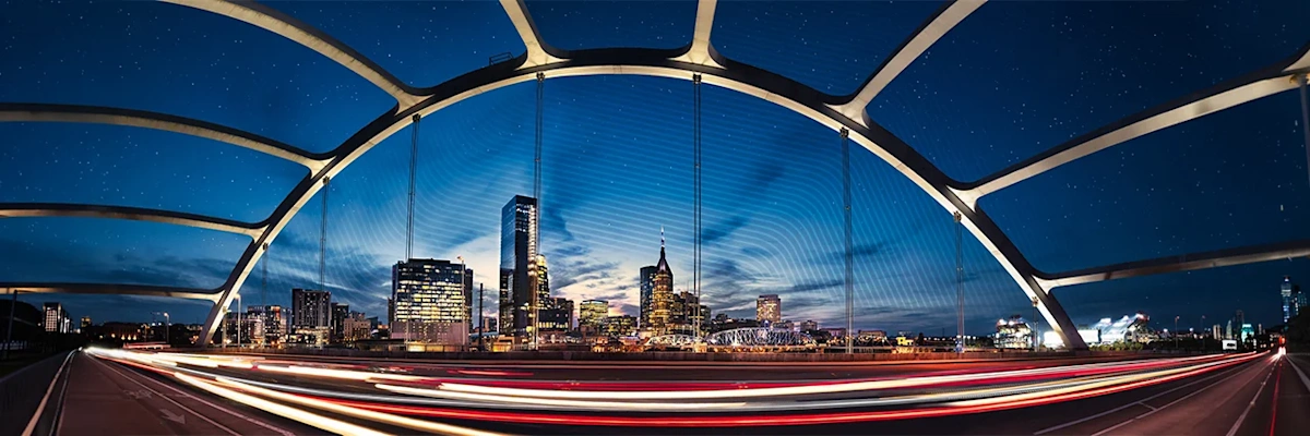 Nashville, TN skyline at night, framed by Korean War Veterans Memorial Bridge. Vehicle light trails pass, highlighting the illuminated AT&T "Batman Building".