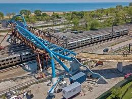 An aerial view of a modern blue pedestrian bridge under construction over train tracks, with a lake and beach visible in the background.