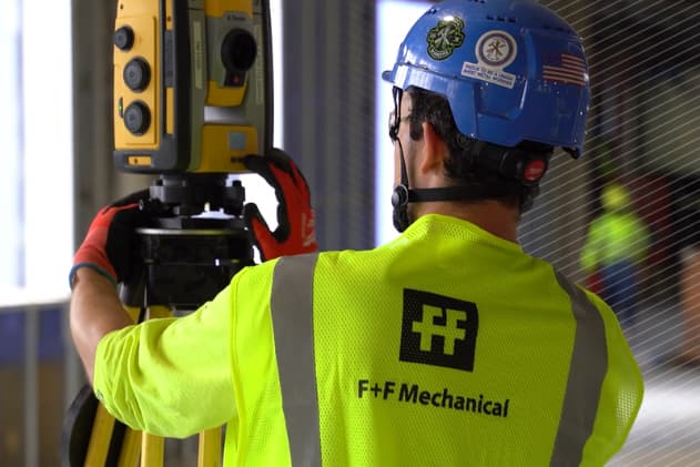 Worker in an F+F Mechanical safety vest operating a Trimble Robotic Total Station at a construction site.
