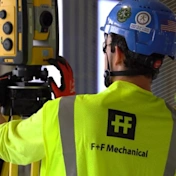 Worker in an F+F Mechanical safety vest operating a Trimble Robotic Total Station at a construction site.