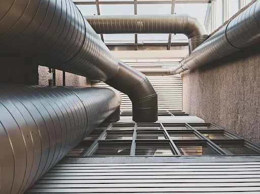 Large, shiny metal HVAC ducts snake across a modern building's interior, featuring a glass ceiling and textured concrete walls.