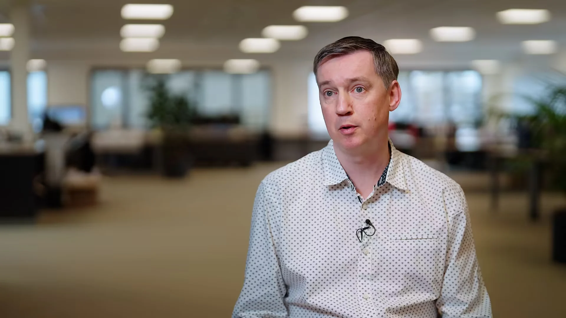 A man wearing a white collared shirt with a small dot pattern is being interviewed in an office setting.