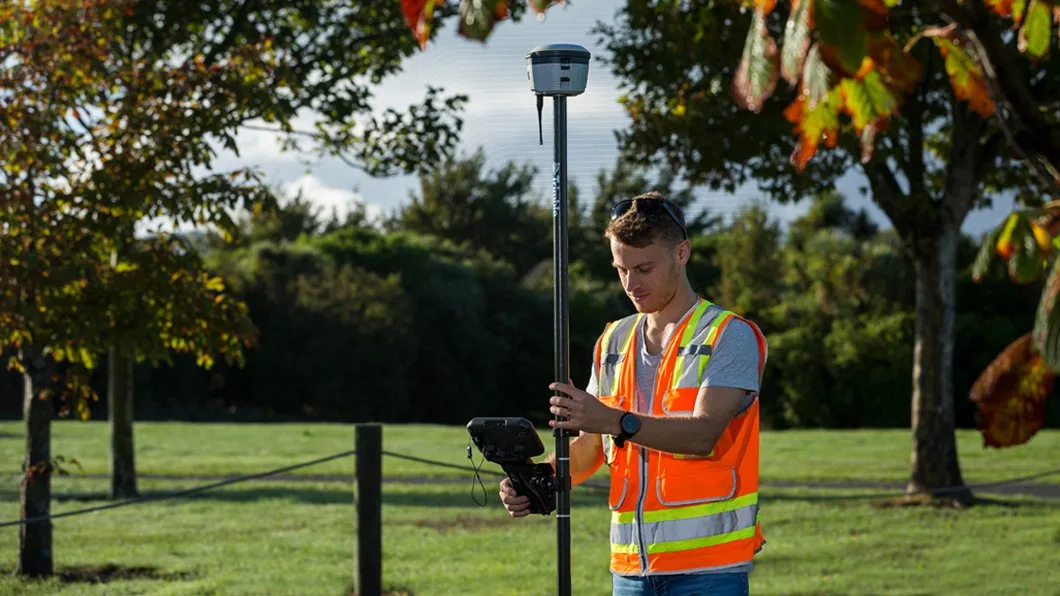 Surveyor in hi-vis vest uses Trimble R10 GNSS receiver on range pole with TSC7 controller for precise site data collection.