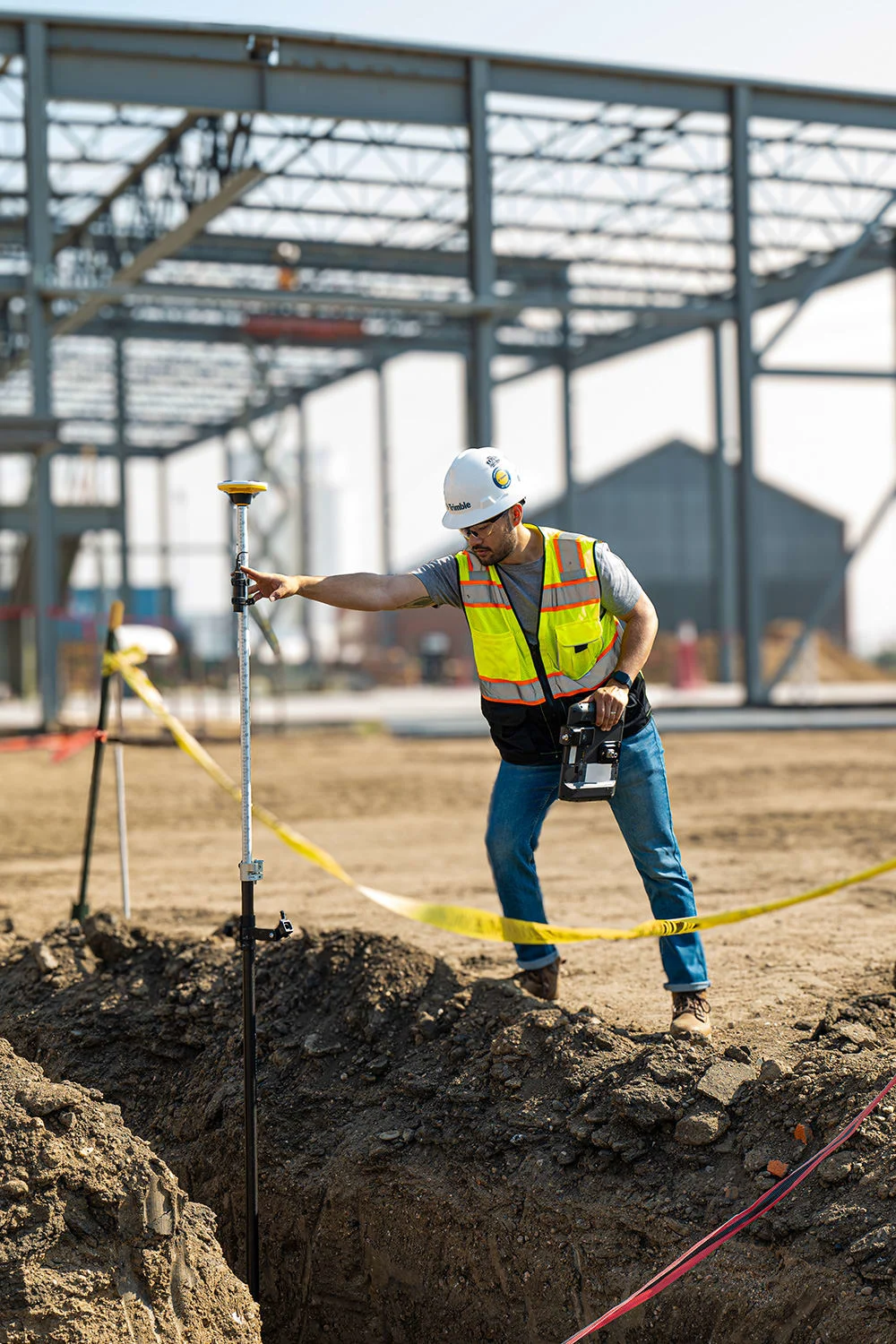 Surveyor using Trimble Catalyst to measure ground height at a construction site with building framing in background.