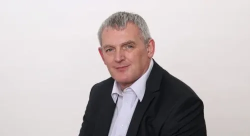 A headshot of a middle-aged man with graying hair, wearing a black suit jacket over a light purple shirt.