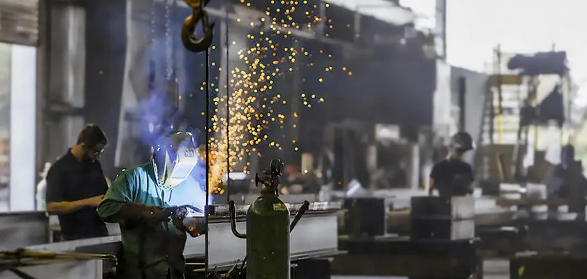 A welder works in a factory, sparks flying, as a crane hovers overhead.