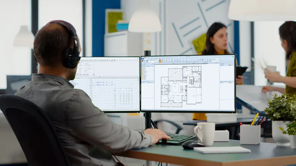 Man wearing headphones, working on architectural blueprints on a computer in a modern office.