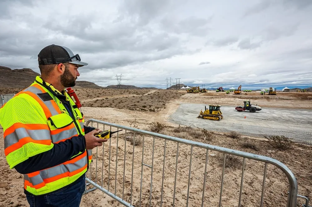 A man in a safety vest holds a remote control, overseeing a construction site with bulldozers and other heavy machinery.