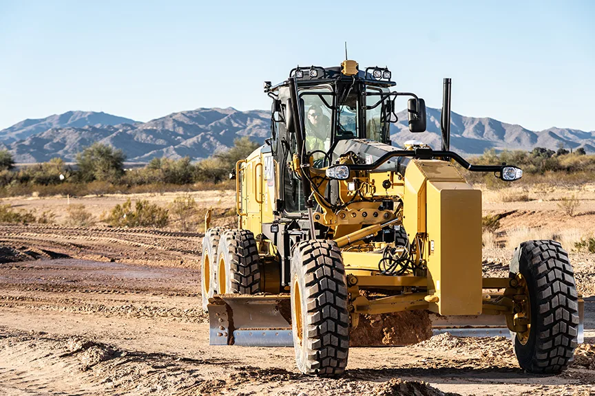 A yellow grader is working on a construction site with mountains in the background.