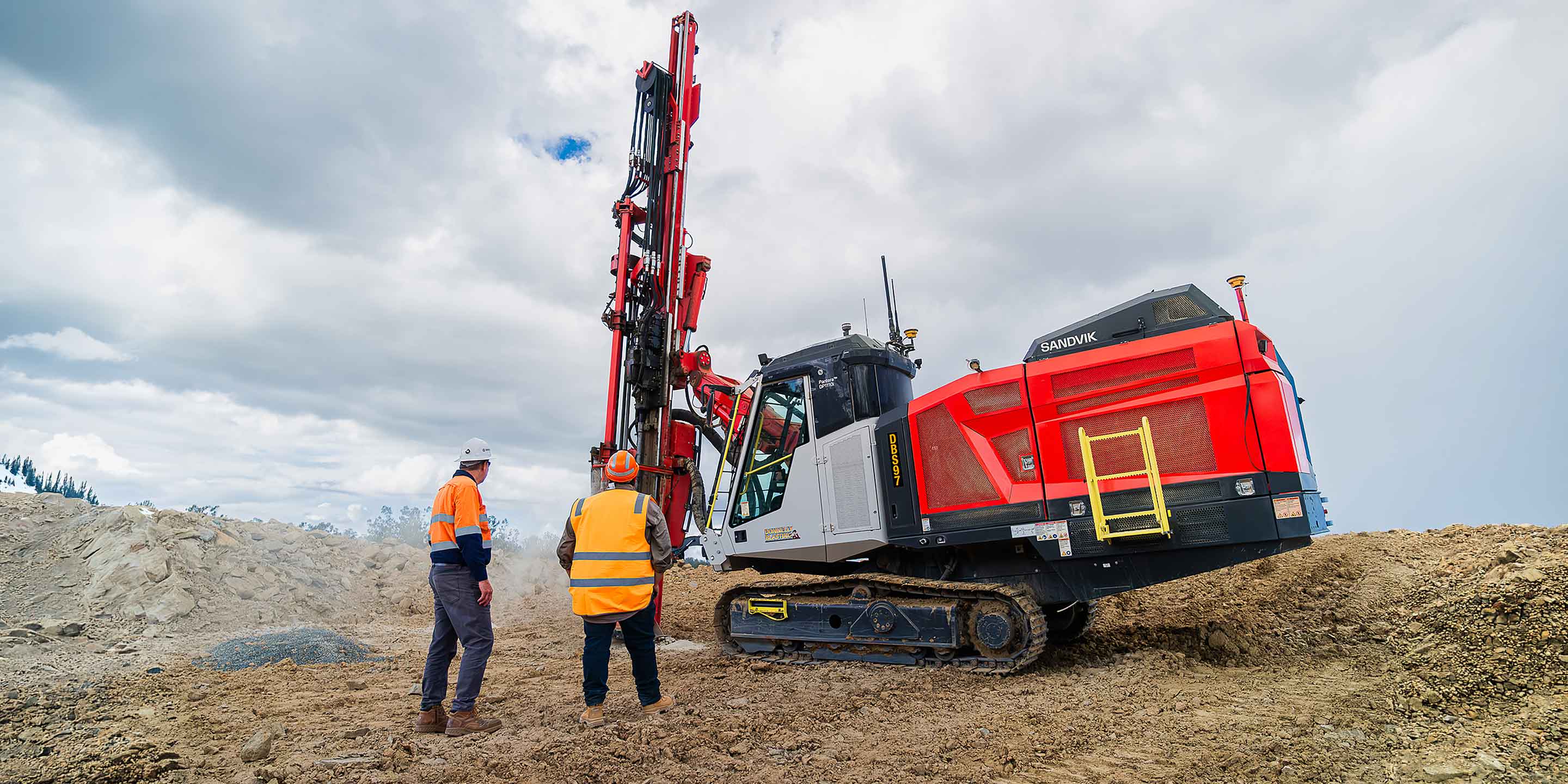Red and white drill rig operating at a quarry site with two workers in high-visibility safety gear standing nearby.