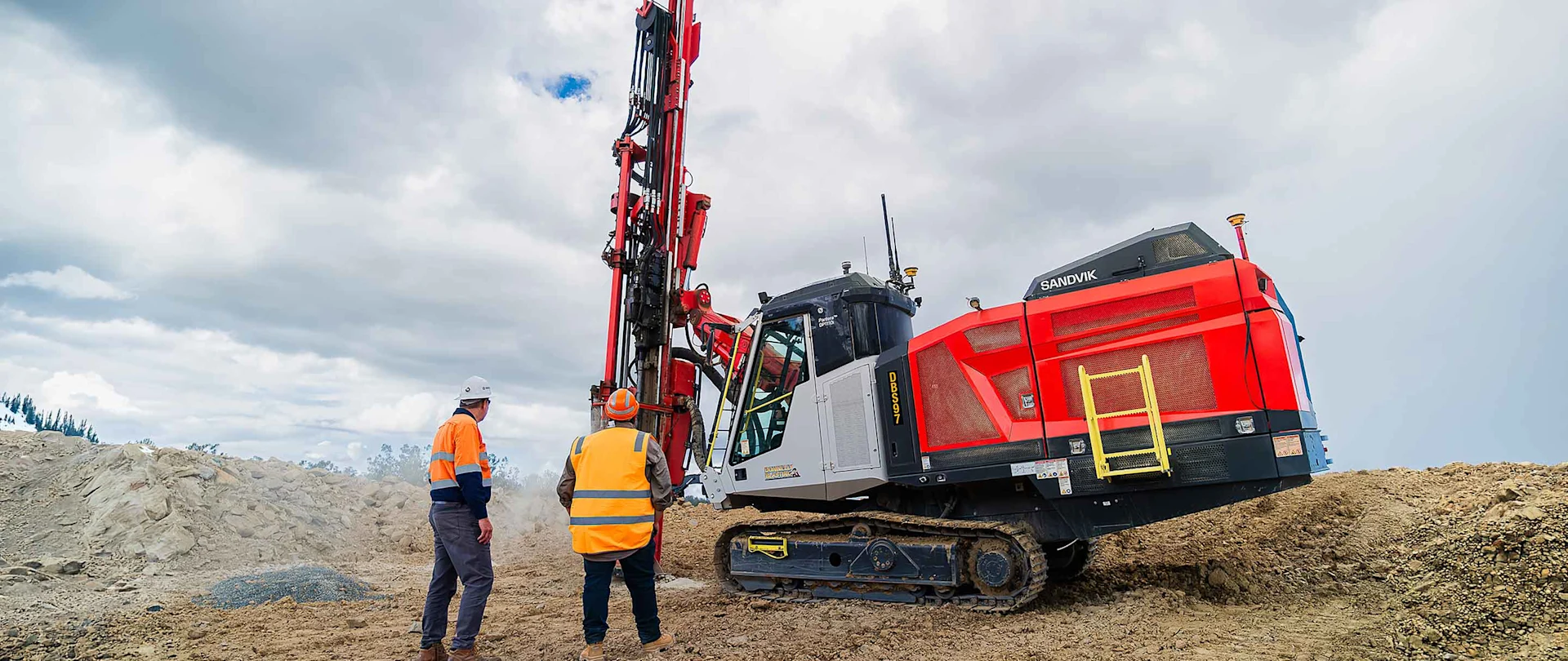 Red and white drill rig operating at a quarry site with two workers in high-visibility safety gear standing nearby.