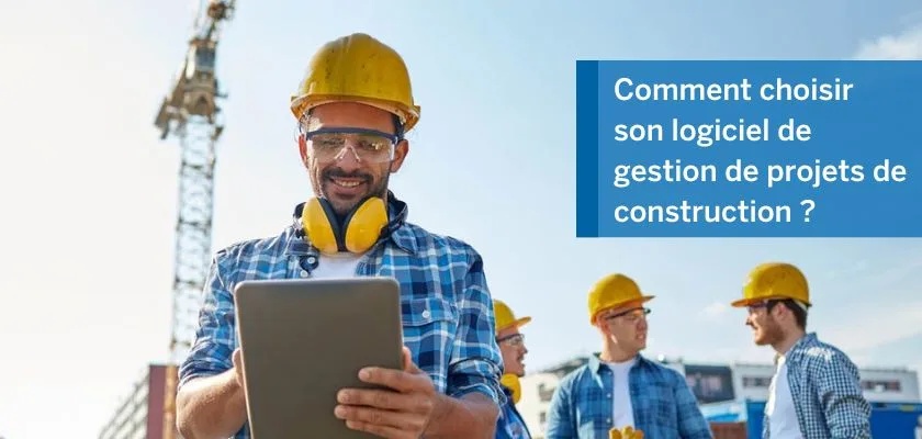 A man in a hard hat and safety glasses uses a tablet while other construction workers stand nearby, with a crane in the background.