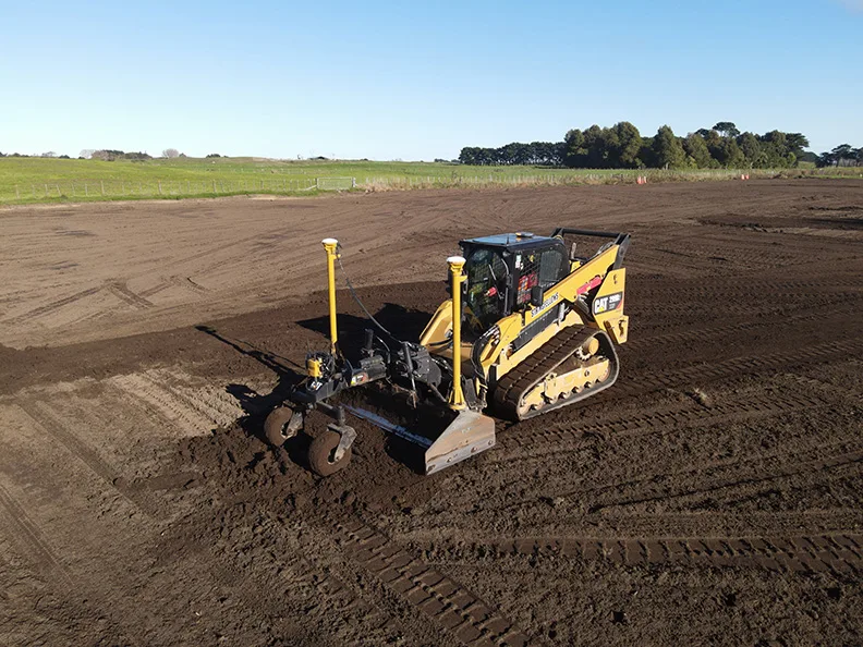 Yellow bulldozer with a grading attachment levels dirt on a construction site under a clear blue sky.