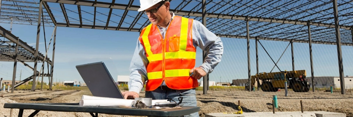 Construction worker in safety gear using a laptop at a site with steel framework overhead