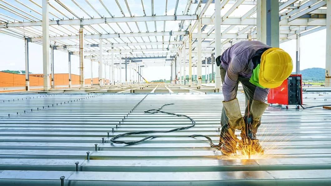 Worker in yellow hard hat welding on metal decking, creating sparks inside an open steel-frame building.