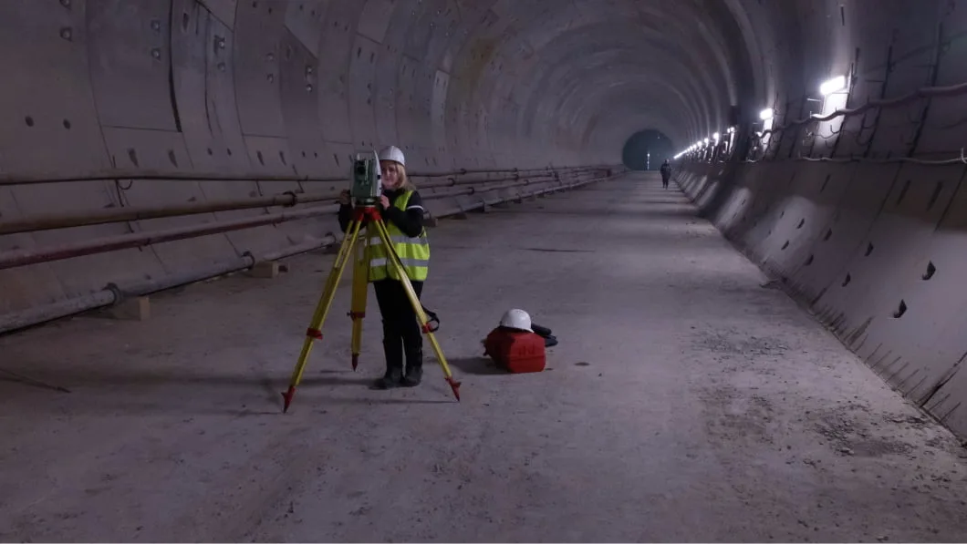A woman in safety gear operates a surveying instrument inside a large, arched concrete tunnel.