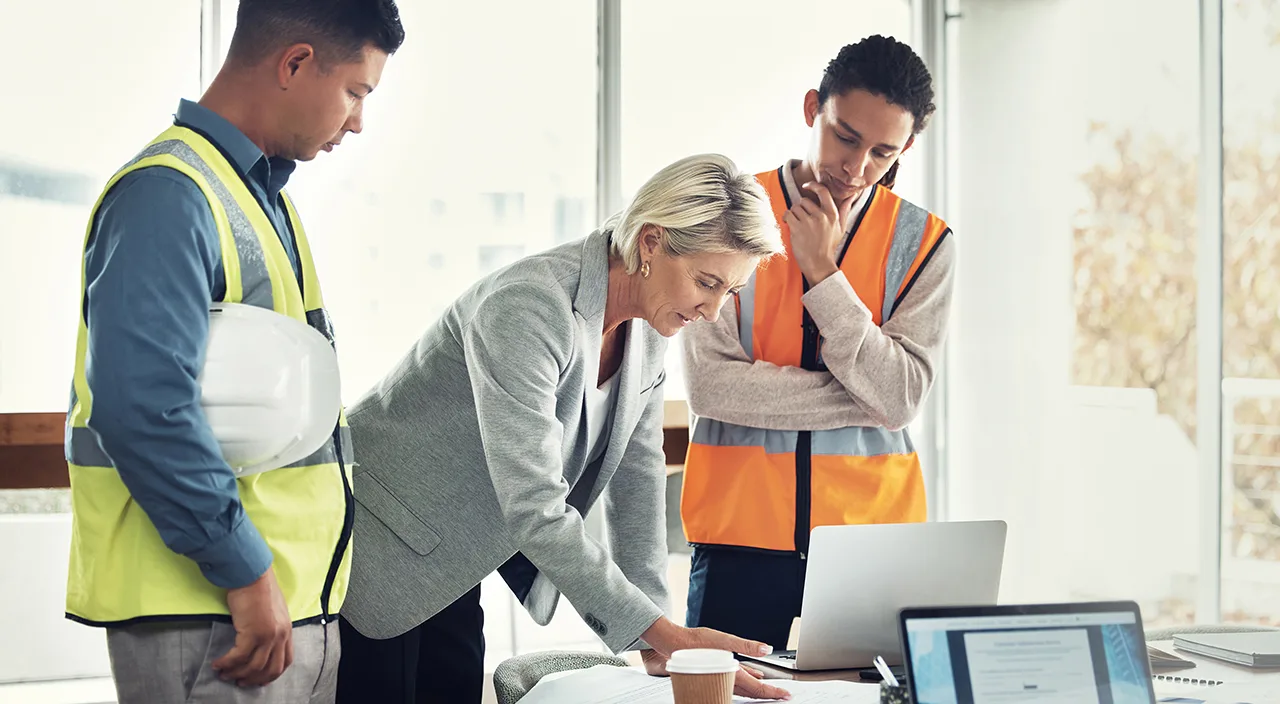 Construction workers and a manager review plans and laptops at a desk in a bright room.