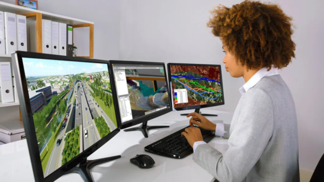 A person sits at a desk typing on a keyboard in front of three computer monitors displaying images and data about roadways.