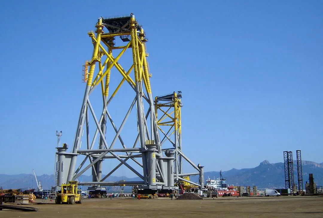 A large yellow and gray offshore jacket structure is being prepared for loadout on a clear day.