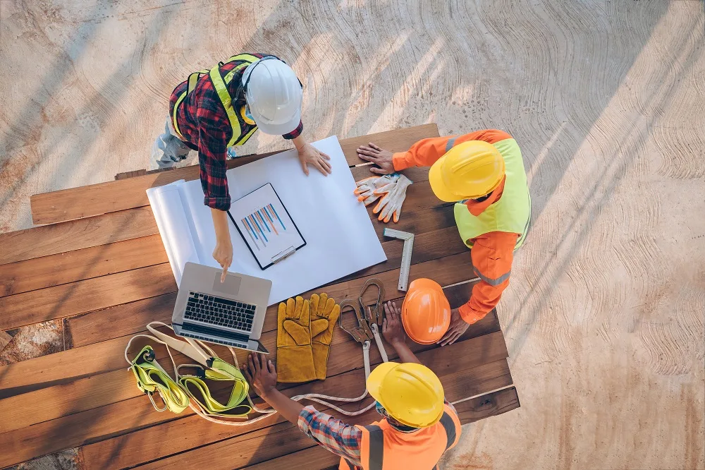 Three construction workers wearing hard hats and safety vests collaborate around a table with blueprints, a laptop, and safety equipment.