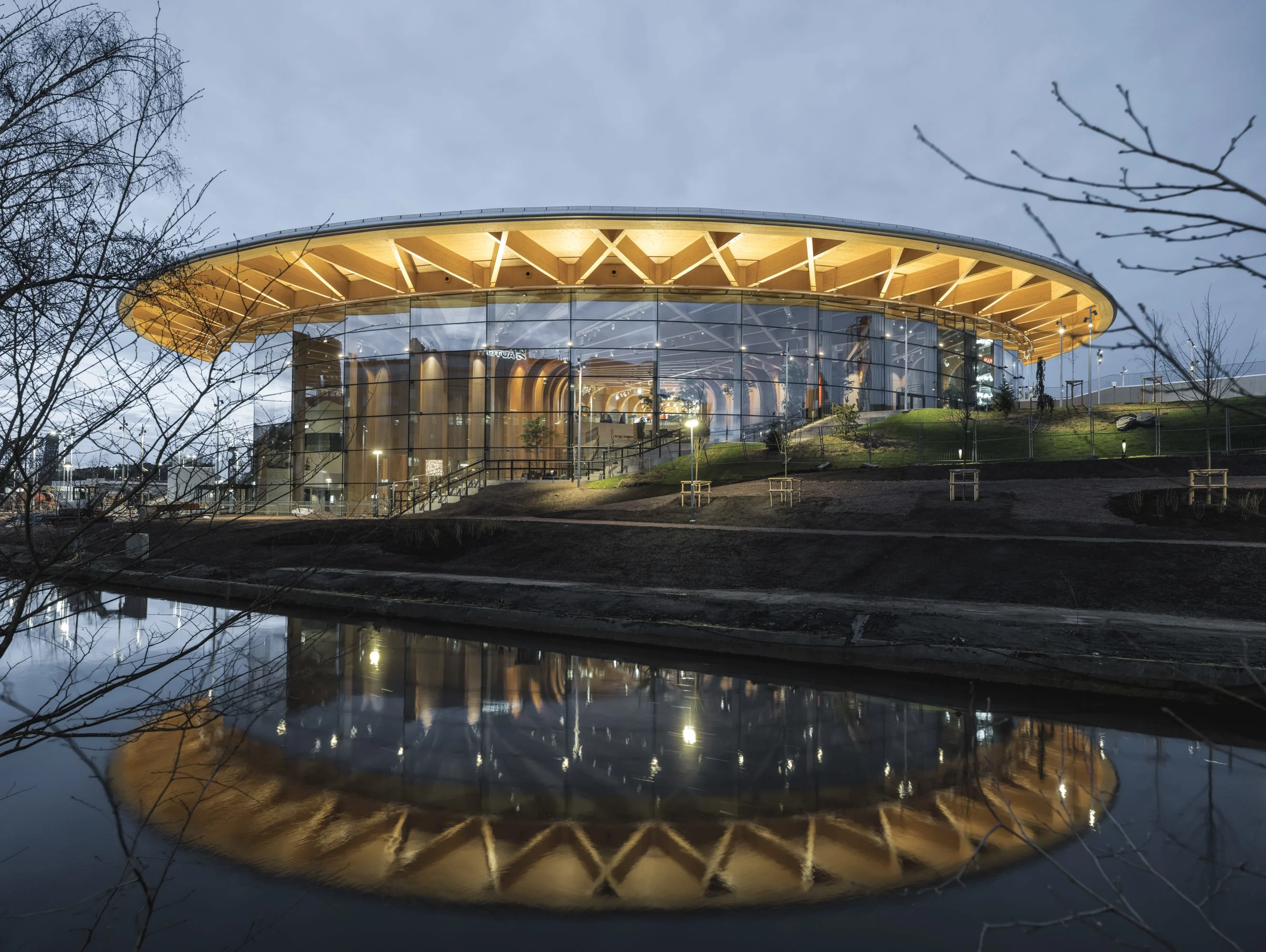 World of Volvo, a modern building with a glass facade and wooden roof, reflected in water at dusk.