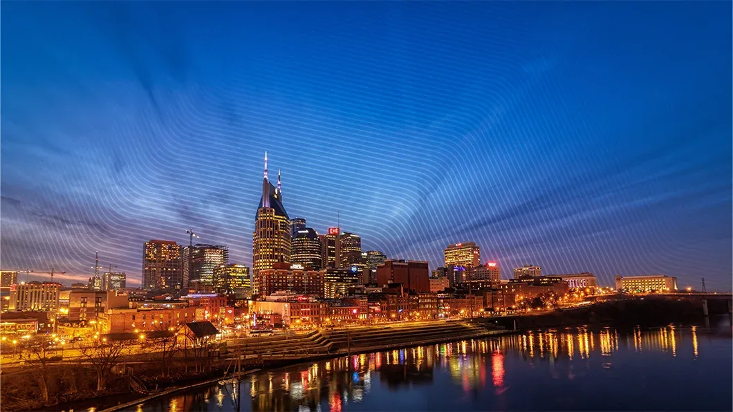 Nashville skyline at dusk with the AT&T "Batman" Building, city lights reflecting on the Cumberland River, and active construction cranes.