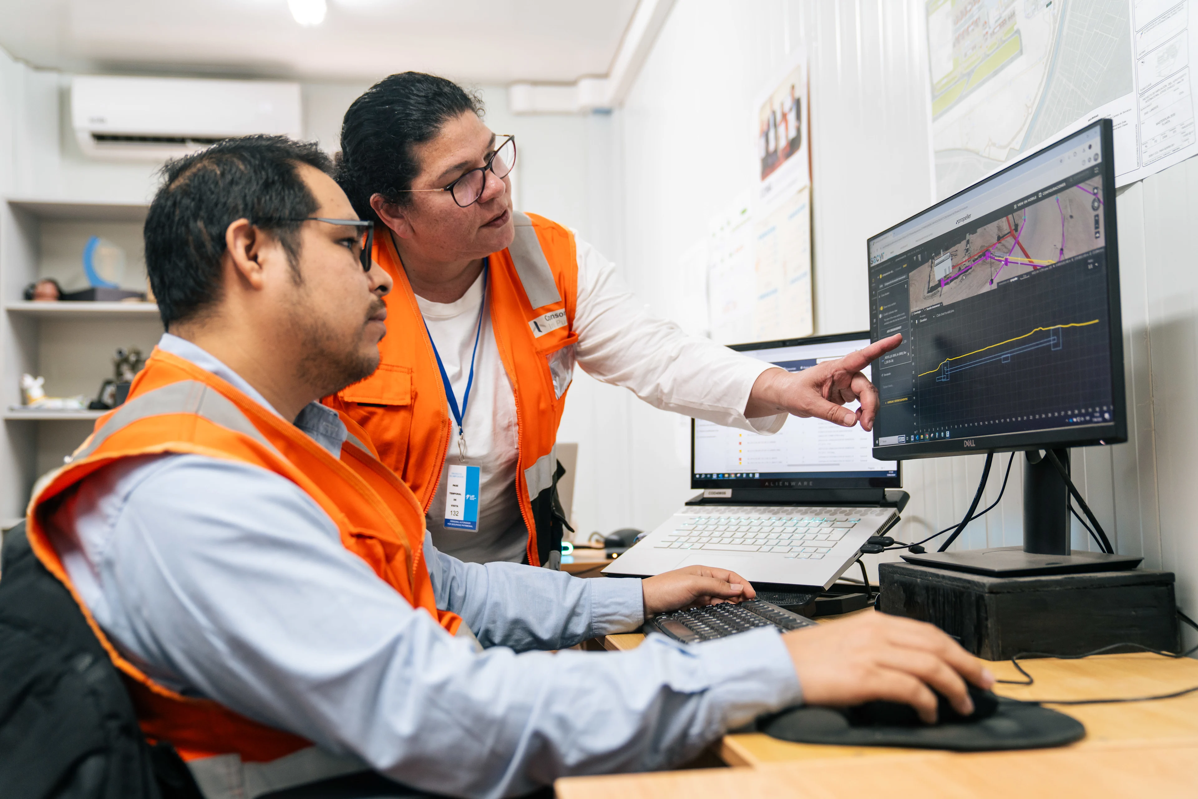 Two engineers in orange safety vests analyze drone survey data on a computer screen, one pointing at the display.