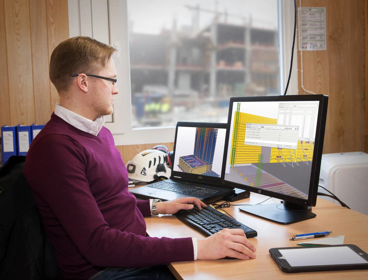 An engineer wearing glasses and a maroon sweater sits at a desk, looking at a computer screen displaying a 3D building model.