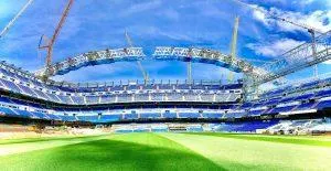 A wide shot of the Santiago Bernabéu stadium with blue seating and a bright green pitch, under construction with cranes.