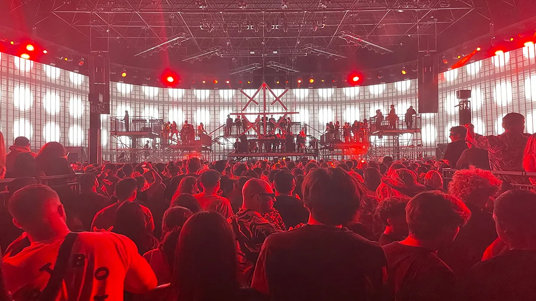 An in-the-crowd perspective looking towards the stage, where red stage lights cast an intense glow over a large group of concertgoers.