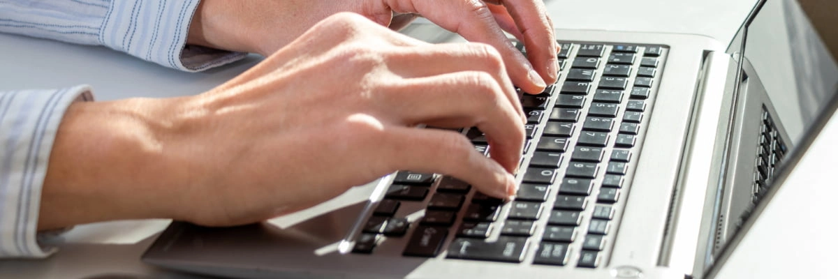 Close-up of a person's hands typing on a laptop keyboard on a white desk.
