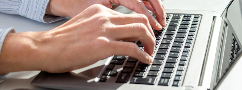 Close-up of a person's hands typing on a laptop keyboard on a white desk.