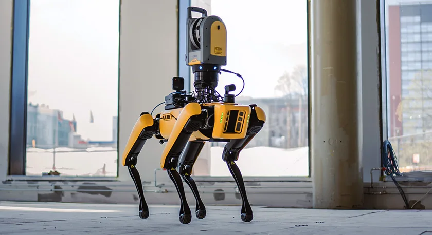 A yellow and black robotic dog with a scanner on its back stands on a concrete floor in a large, unfinished room.