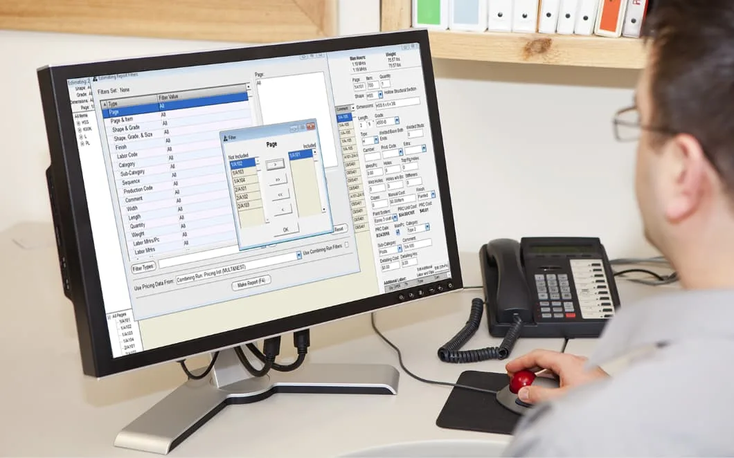 A man in glasses uses a computer to input data on a desktop with a phone and trackball mouse.