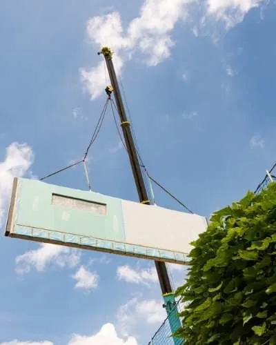 A modular wall panel is lifted into place by a crane against a bright blue sky with fluffy white clouds.