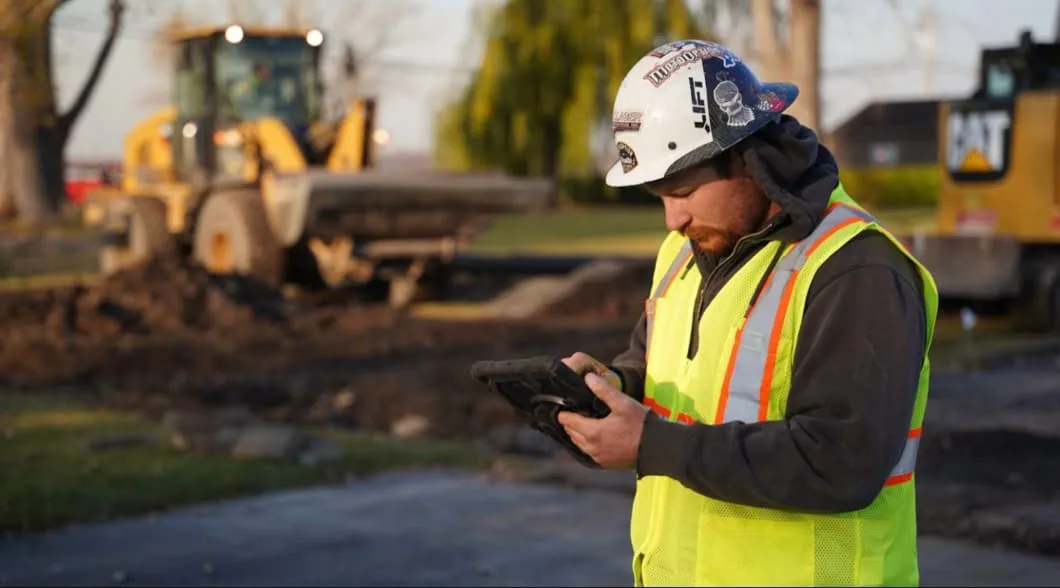 A construction worker in a yellow safety vest and hard hat looks at a tablet while a bulldozer works in the background.