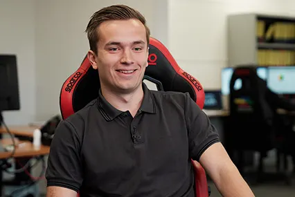 Person smiling in a black polo shirt seated in a red gaming chair in an office environment with computers visible.