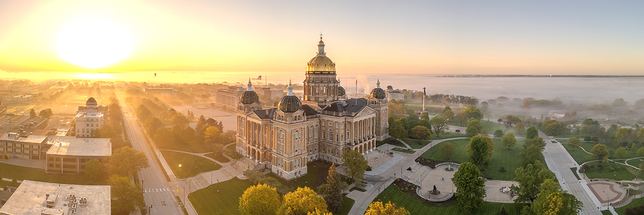 The Iowa State Capitol at sunrise, its Renaissance sandstone and brick architecture, featuring a gold-leaf central dome and four other domes, emerges from fog.