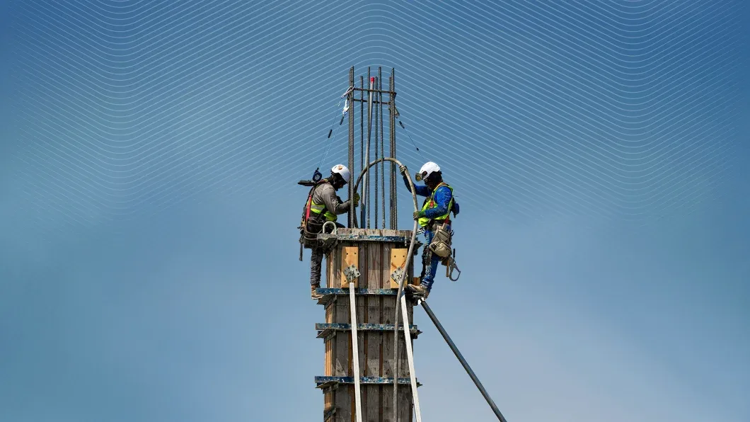 Two construction workers in safety gear work atop a tall concrete column formwork against a blue sky with stylized white wavy lines.