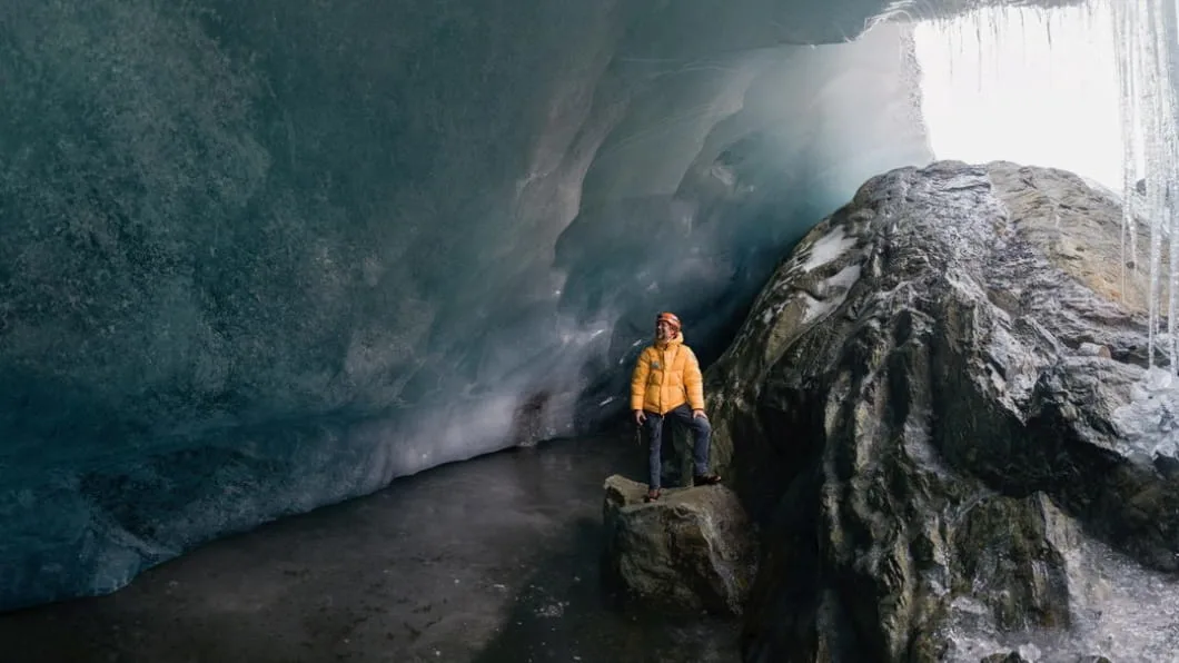 Klaus Thymann in a yellow jacket standing on a rock inside a vast ice cave with icicles at the entrance.