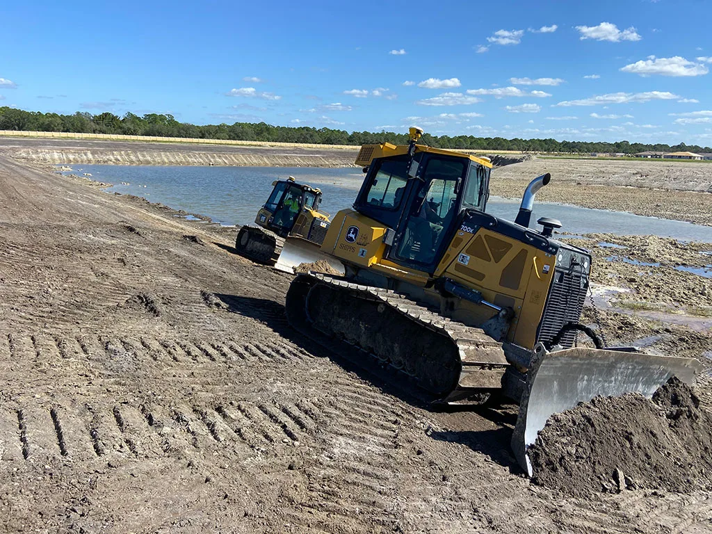 Two yellow John Deere bulldozers work on a muddy construction site, moving dirt and water under a blue sky.