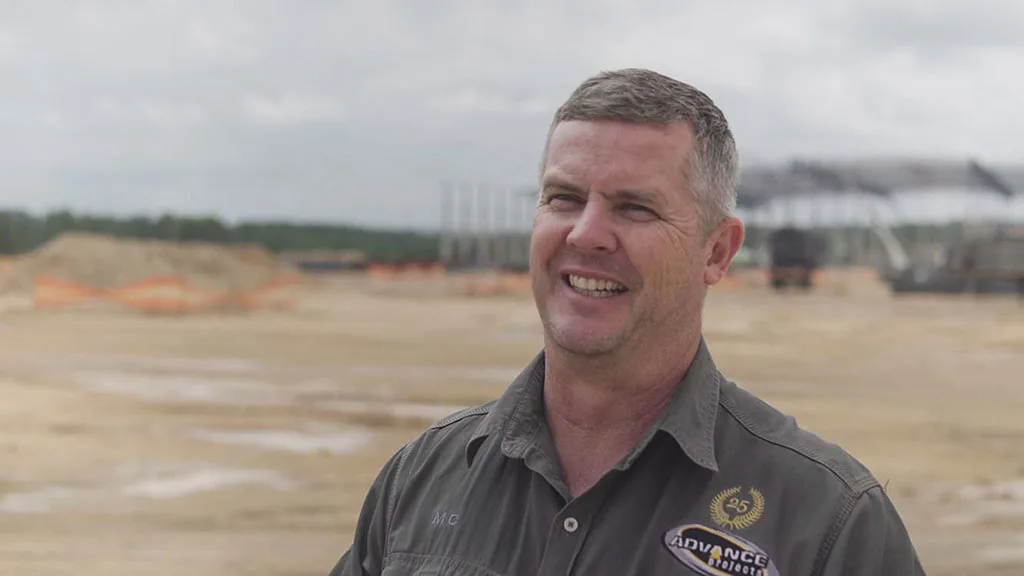 Smiling man in an "ADVANCE Projects" work shirt at a construction site.