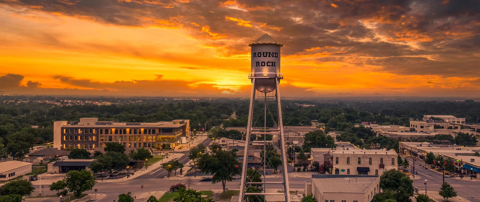 A water tower labeled Round Rock at sunset, with orange and yellow clouds in the sky and greenery and buildings below.