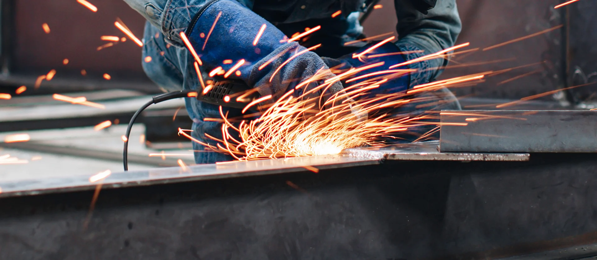 Close-up shot of a metal fabricator actively grinding or welding structural steel, creating a bright, dense shower of orange sparks across the industrial workshop floor.
