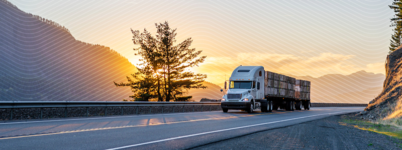 Semi-truck driving on a scenic highway at sunset, with mountains and trees in the background.