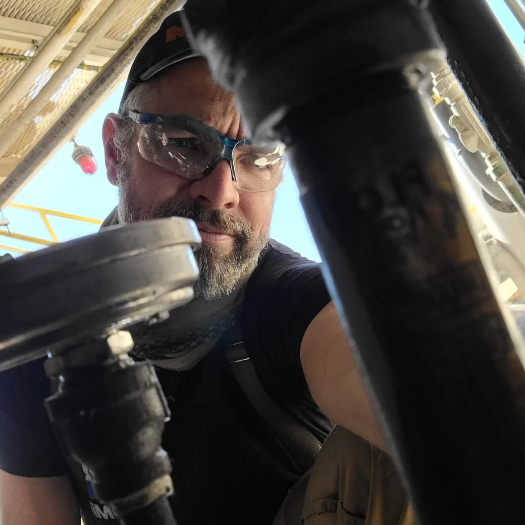 A man wearing safety glasses and a black cap looks down at pipes.