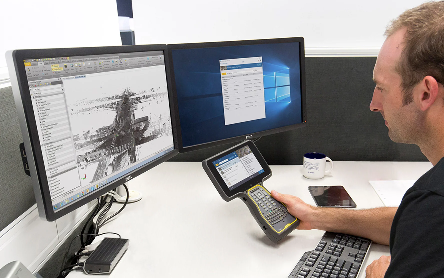 Man reviews 3D scan data on dual monitors and handheld device at desk.